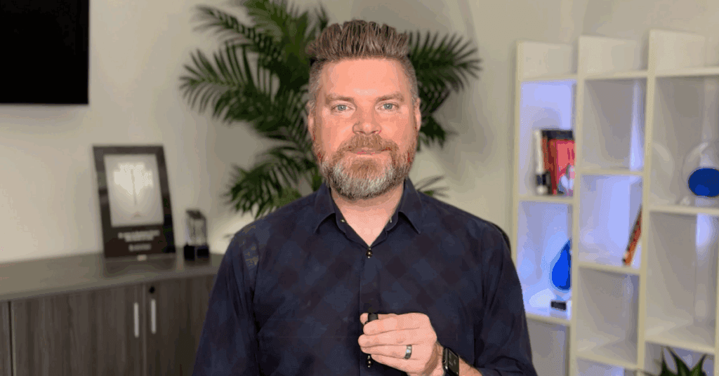 Andrew Aebersold, CEO of Mediaura, looks into the camera while explaining what to avoid when hiring a digital marketer. He is wearing a dark blue checkerboard print collared shirt. The background is an office scene, featuring a bookshelf and plant.